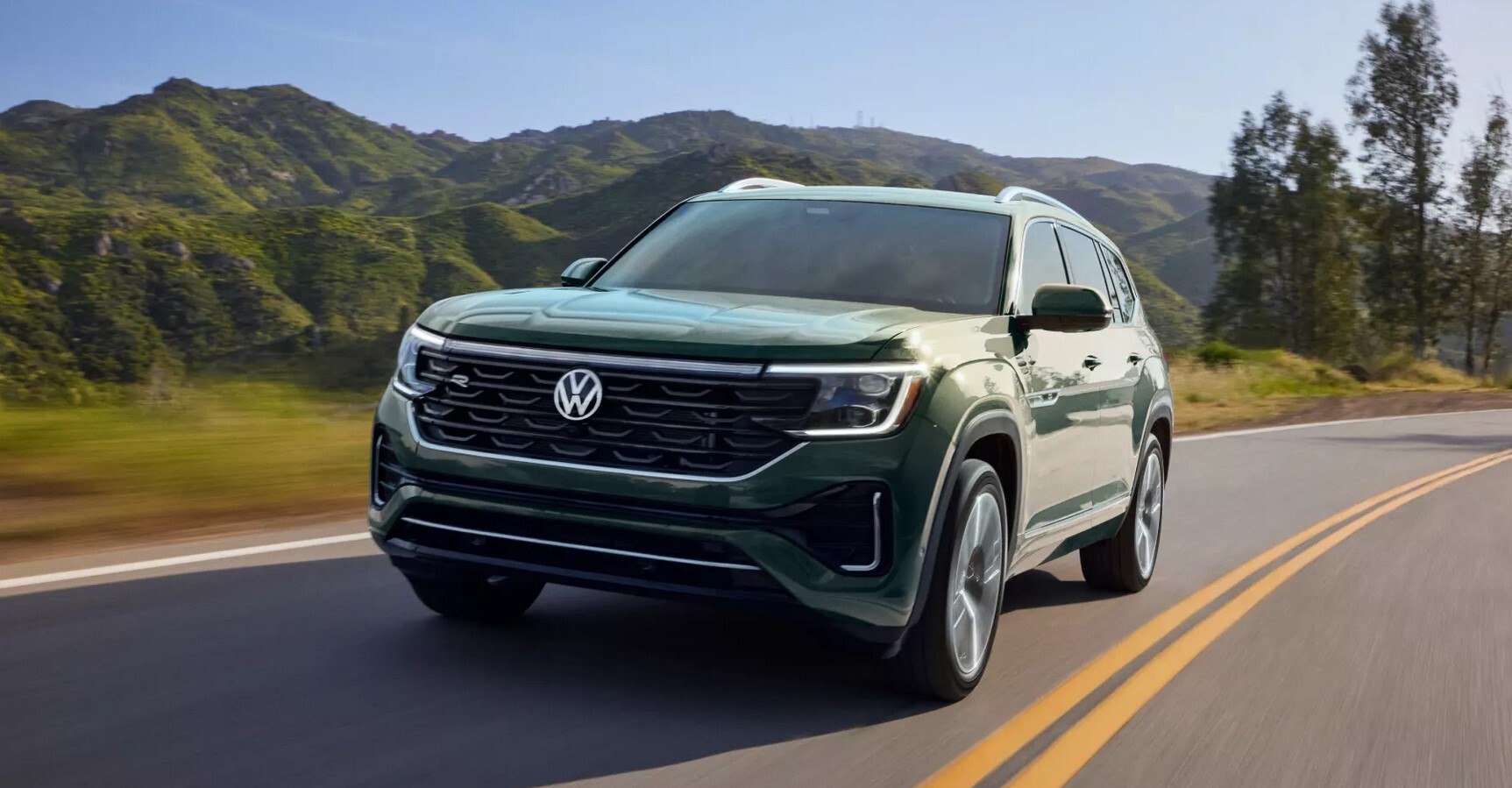 A green Volkswagen Atlas drives on a winding road with mountains in the background