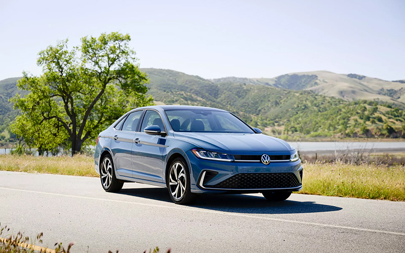 A blue Volkswagen Jetta parked on a scenic road with rolling hills in the background.