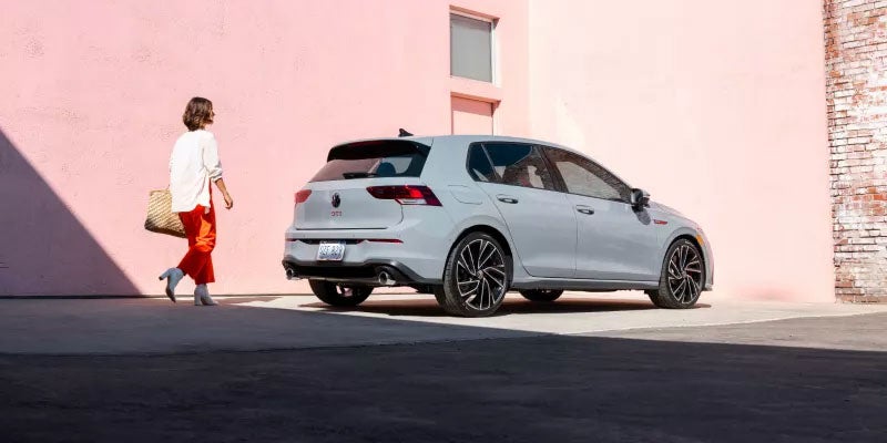 Gray Volkswagen GTI hatchback parked near a pink building with a person walking nearby.